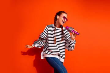 Joyful young woman with red hair sings into pink microphone against vibrant orange background