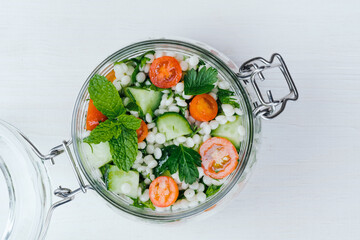 Close up fresh couscous salad in a glass jar with cucumbers, tomatoes, herbs, lemon on a white...