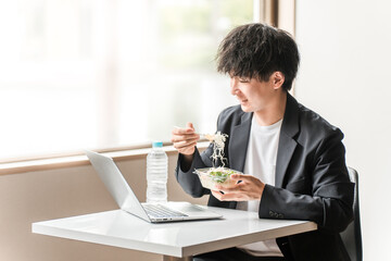 イートインスペース・社員食堂でパソコンを見ながら昼食にお弁当を食べる若いアジア人男性ビジネスマン

