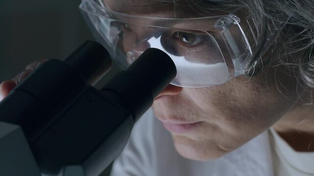 Focused female scientist, wearing protective eyewear, looking through microscope while examining specimen in laboratory. Close-up view