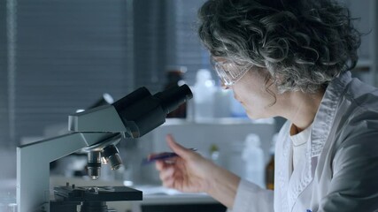 Middle-aged female scientist with curly grey hair, wearing protective goggles, observing specimen under microscope and making notes during research in laboratory