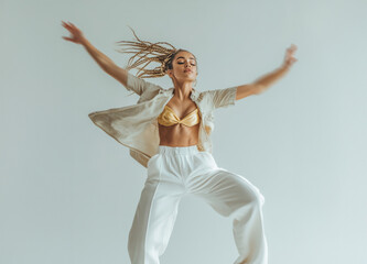 Fototapeta premium Young woman in fashionable, light toned hip hop and performance dance outfit performs high energy dance move against white studio background, radiating confidence and strength.