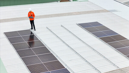 A technician in a hard hat and high visibility jacket inspects solar panels on a large metal rooftop. The aerial view emphasizes renewable energy, solar maintenance, and sustainable infrastructure.