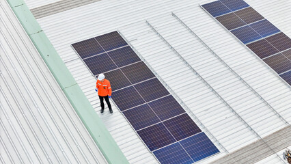 A technician in a hard hat and high visibility jacket inspects solar panels on a large metal rooftop. The aerial view emphasizes renewable energy, solar maintenance, and sustainable infrastructure.