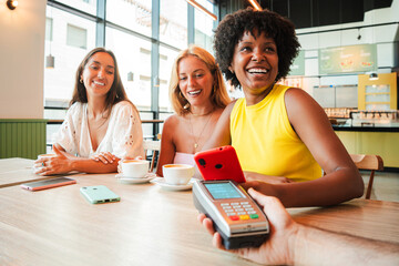 three diverse happy young women friends smiling while making contactless mobile payment with...