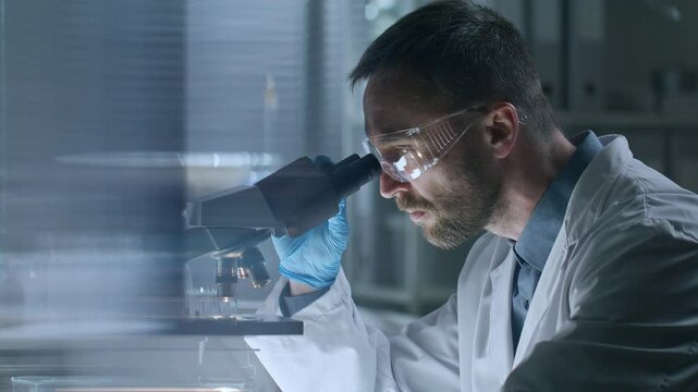 Scientist wearing safety goggles, gloves and lab coat, placing glass with sample under microscope and examining it in laboratory. Side view