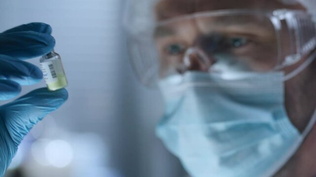 Lab scientist in protective face mask, goggles, disposable cap and gloves studying yellow substance in glass vial during medical test or research. Close-up, rack focus shot