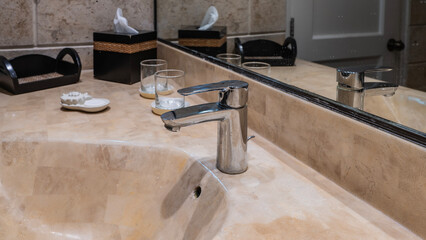 A metal faucet on the edge of a beige wash basin. A glass, a soap dish, and a box with napkins next to it on the countertop. Reflection in the wall mirror.