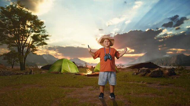 Asian Boy With A Hat And Binoculars Using The Magnifying Glass Then Flexing His Bicep While Tent Camp Lakeside. Boy Researcher Examines Something, Travel Tourism Adventure, Full Body