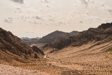 A winding road through a desolate desert valley with rocky hillsides and a cloudy sky.
