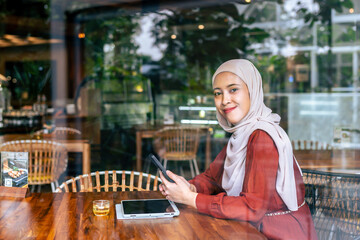 Confident young woman in a cafe uses her smartphone and tablet. She wears a hijab, creating a blend of traditional and modern elements, showcasing connectivity and relaxation.