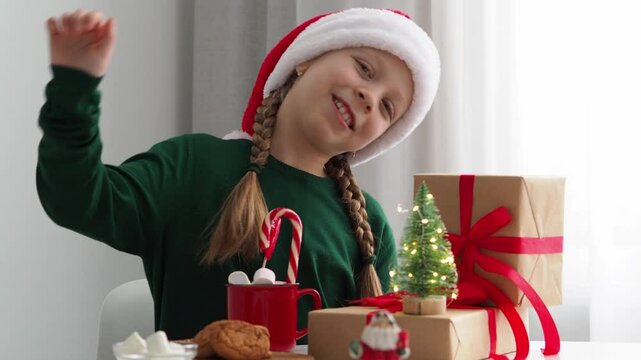 A happy Girl in a green jumper and a red Santa hat, sitting at a table, sings New Year and Christmas songs and dances. Festive decor on the table. A gift box. A happy child on Christmas Eve.