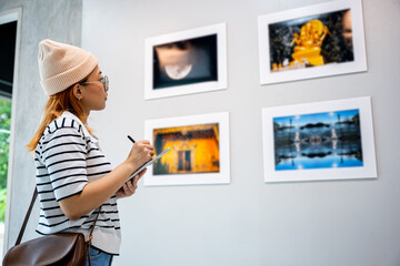 Asian woman note information to tablet at art gallery collection in front framed paintings looking pictures on wall, Young person at photo frame write digital book at show exhibit artwork gallery © sorapop