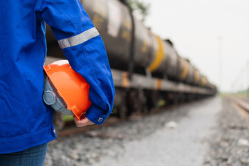 A worker in blue coverall uniform is holding an orange safety hardhat or protective helmet, standing in front of crude oil train tanker. Ready to work in challenge industrial scene, close-up. 