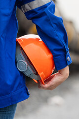 A worker in blue coverall uniform is holding an orange safety hardhat or protective helmet,...