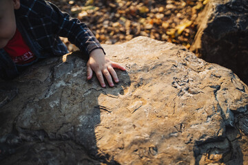 A young boy is seated comfortably on top of a large rock while resting his hands on its surface, relishing the tranquil moment amidst the stunning beauty of nature surrounding him