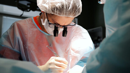 Skilled surgeon in surgical attire, including a mask and loupes, carefully performs a surgical...