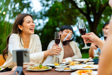 Group of Happy Asian millennials people enjoy and fun celebration reunion meeting using mobile phone video calling talking with friends at outdoor dinner party at restaurant on holiday vacation.