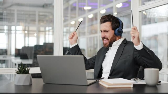 Businessman having fun listening to music, using pens as drumsticks, playing air drums in office. Man enjoys music, relaxing and having fun at work.