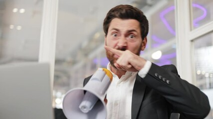 Angry businessman yells into a megaphone and points his finger at camera. Man is sitting at desk in office environment. Annoyed male in formal clothes speaking in megaphone and making hand gestures. .