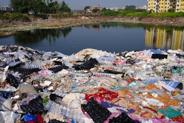 A large pile of discarded textiles and waste materials accumulating near a body of water likely...