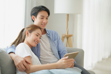 Asian young couple watch movie online by tablet on sofa, leisure spending weekend time together at home. Attractive Husband with Wife with positive emotion lover.