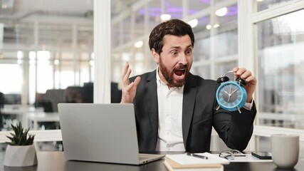 Stressed businessman holds blue alarm clock and shouts in despair while sitting at desk in office. This video portrays themes of business, stress, and time management.