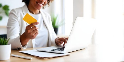 Beautiful woman in white blouse, sitting at her bright home at her desk and holding a credit card with her hand while making an online purchase on her laptop