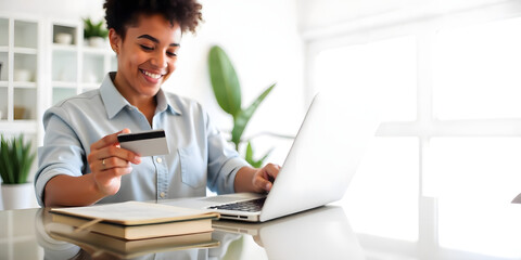 Beautiful woman in white blouse, sitting at her bright home at her desk and holding a credit card with her hand while making an online purchase on her laptop