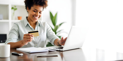 Beautiful woman in white blouse, sitting at her bright home at her desk and holding a credit card with her hand while making an online purchase on her laptop