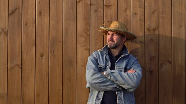 A man in a straw hat stands against a wooden wall. Dressed in a denim jacket. Bearded. Looking into the distance. Warm clothes. Farmer. Working man. Wooden boards in the background. Close-up. Serious 
