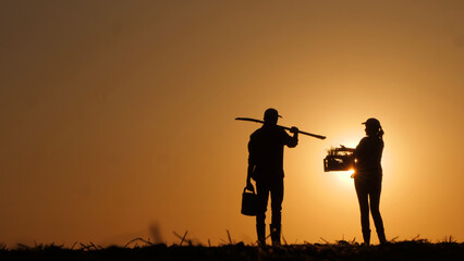 Against the backdrop of the setting sun, silhouettes of a man and a woman farmer can be seen in the field.