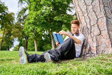 A man sits on the grass in a park, leaning against a tree while absorbed in reading a book,...
