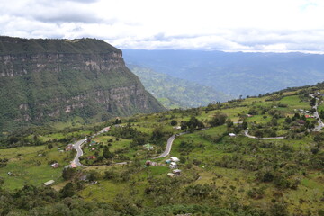 Naklejka premium En esta imagen se observa un paisaje montañoso con una carretera serpenteante que atraviesa una zona rural. Hay varias áreas verdes y pequeñas construcciones dispersas, probablemente casas o edificaci