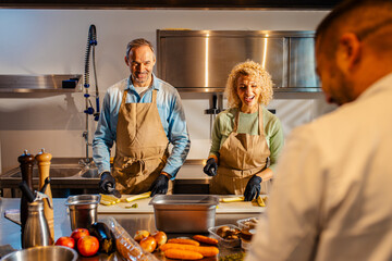 Middle-aged couple enjoying a cooking class with a chef preparing zucchini for a meal in the kitchen.
