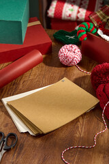 Christmas themed photo with diagonal view from above, showing a batch of blank paper lying on a wooden table, next to wool items, green and red gift boxes, and red paper rolls.