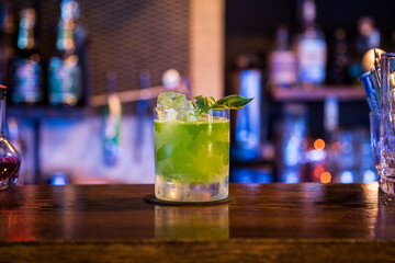 A bright green cocktail sits on a wooden bar, garnished with basil leaves and ice cubes. The background is blurred with soft, colorful bar lights.