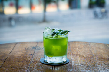 A bright green cocktail sits on a wooden bar, garnished with basil leaves and ice cubes. The background is blurred with soft, colorful bar lights.