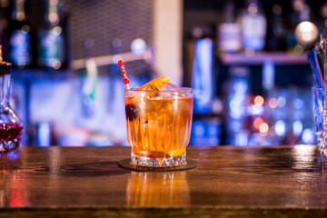An amber cocktail in a glass with ice, garnished with an orange slice and a red stirrer, sits on a bar with blurred lights in the background.