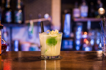 A cocktail stands on a wooden table in the background colorful lights can be seen