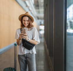Young happy woman traveler in hat with suitcase at airport or train station waiting for travel