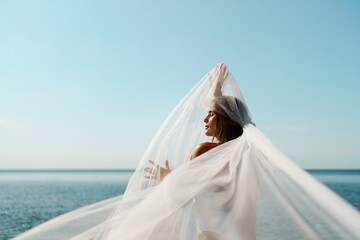 A woman stands on a beach with a white veil over her head. The scene is serene and peaceful, with...