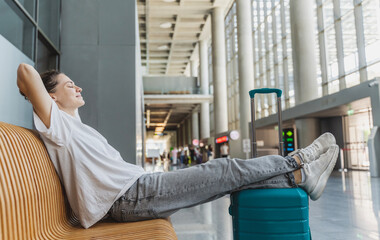 Young happy woman traveler relaxing on bench waiting for flight with suitcase in departure area at...
