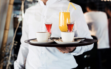 Waiter holding a tray with tea, coffee, and juice in a cozy cafe setting, showcasing professional service and a variety of beverage options for customers in a welcoming, relaxed atmosphere.