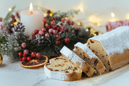 Traditional German Christmas cake Stollen with raisins, fruits and nuts inside and sprinkled with powdered sugar on a festive table decorated with Christmas tree branch, lights and candles.