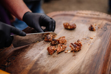 A close-up of a person wearing black gloves slicing grilled meat pieces on a wooden board, highlighting texture and preparation in outdoor cooking..