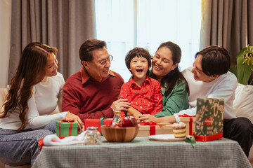 parent, parenthood, family, celebrate, xmas, present, celebration, laughing, christmas, brother. A family of five is sitting around a table. The family is smiling and enjoying each other's company.