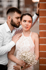 Pretty bride in a tight dress stands leaning against a brick wall while her groom leans his nose to her ear