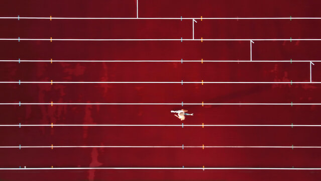 Aerial view of athlete on red track field. A lone athlete runs on a striking red track, captured from above, with white lane markings and colorful indicators.