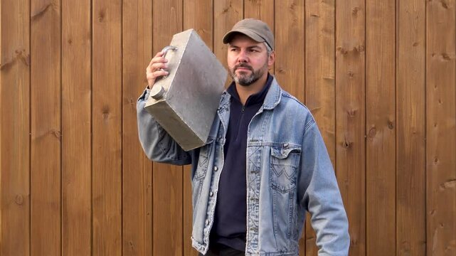 A man with an iron canister. Passing a wooden wall. Dressed warmly. Bearded. Denim jacket and baseball cap. Rural area.